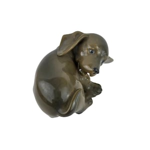 May include: A ceramic figurine of a dachshund puppy in a curled-up position. The dog is a muted olive green color with a glossy finish. The puppy's ears are floppy, and its nose and eyes are black.
