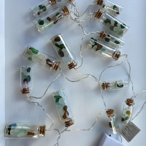May include: Fairy lights with small glass bottles filled with colourful sea glass. Each bottle has a cork stopper and is connected by a thin silver wire. A white battery pack is also visible.