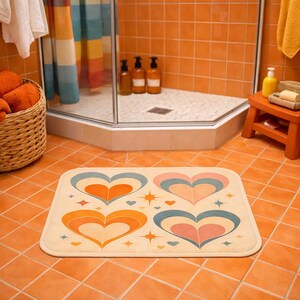 May include: A rectangular bath mat with a cream background and four retro-style hearts in orange, blue, and pink. Small starburst designs are scattered around the hearts. The mat is in a bathroom with orange tile.