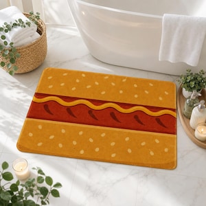 May include: A rectangular bath mat featuring a graphic print of a hamburger bun with sesame seeds, a red layer representing ketchup, and a wavy yellow line representing mustard, designed with a flat, non-textured surface finish.
