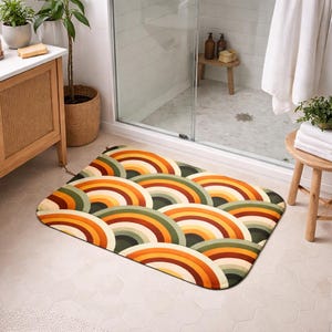 Peut inclure: Un tapis de bain rectangulaire avec un motif arc-en-ciel rétro dans des tons d'orange, de jaune, de vert et de marron. Le tapis est posé sur un sol carrelé blanc dans une salle de bain. Le motif présente des demi-cercles superposés.
