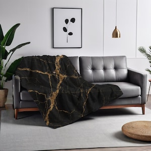Op de afbeelding: Een zwarte en gouden marmer gepatroneerde deken gedrapeerd over een grijze bank. De deken heeft een luxe en moderne uitstraling.