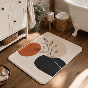 May include: A rectangular bathroom rug with a cream-colored base featuring a printed design of three overlapping abstract shapes in burnt orange, beige, and dark charcoal, overlaid with a delicate black line drawing of a leafy branch.
