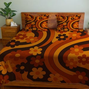 Op de afbeelding: Een beddengoedset in retrostijl met een bloemenpatroon in bruin, oranje en gele tinten. De set bevat een hoeslaken, een laken en twee kussenslopen. Het ontwerp bevat grote, gestileerde bloemen en gebogen lijnen.