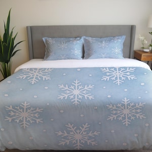 Op de afbeelding: Lichtblauwe beddengoedset met een wit sneeuwvlokkenpatroon. De set bevat een dekbedovertrek en twee kussenslopen. De sneeuwvlokken zijn gelijkmatig verdeeld over het dekbed en de kussens, wat een winterthema creëert.