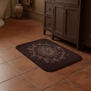May include: A rectangular, dark brown bath mat with a floral design in a lighter brown color. The floral design is centered and surrounded by a leafy vine pattern. The mat is placed on a brown tile floor.