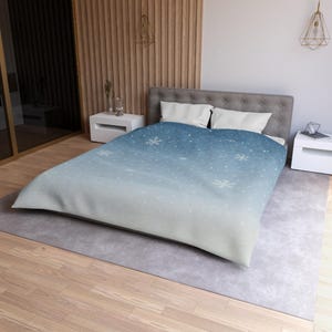May include: A blue and white duvet cover with a winter theme. The design features a gradient of blue and white, with scattered snowflakes. The bedding is on a bed with white pillows and a gray headboard.
