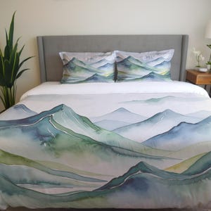 以下が含まれることがあります： 緑、青、グレーの色合いの水彩画の山岳風景のデザインが施された白い羽毛布団カバー。羽毛布団カバーは、2つの同じ枕カバーが付いたベッドの上に置かれています。