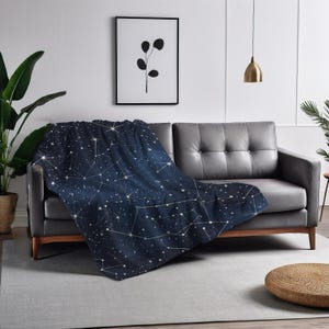 May include: A navy blue throw blanket with a constellation pattern featuring white stars and lines. The blanket is draped over a gray couch in a living room setting.