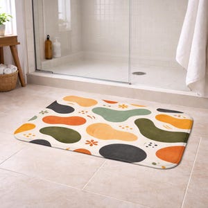 Può includere: Un tappetino da bagno rettangolare con un motivo astratto di forme organiche arancioni, verdi, nere e gialle su sfondo crema. Piccoli dettagli di fiori e punti sono sparsi. Il tappetino è in un bagno.