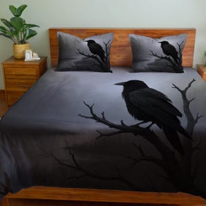 Puede incluir: Un juego de cama con un diseño oscuro. La funda nórdica y las dos fundas de almohada muestran un cuervo negro posado en una rama de árbol desnuda sobre un fondo gris brumoso. El tema general es gótico e inspirado en la naturaleza.