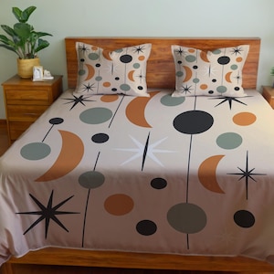 Puede incluir: Un edredón beige y dos fundas de almohada presentan un diseño moderno de mediados de siglo con lunas crecientes naranjas, círculos negros y estrellas. El juego de cama está sobre una estructura de cama de madera.
