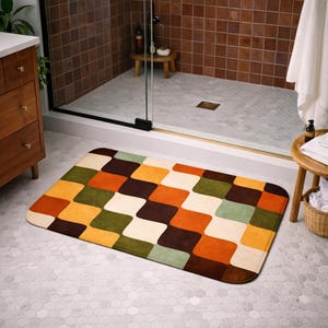 Puede incluir: Una alfombra de baño rectangular con un diseño retro. La alfombra presenta un patrón repetitivo de cuadrados redondeados en tonos de marrón, naranja, amarillo, verde y crema. La alfombra está colocada sobre un suelo embaldosado frente a una ducha.