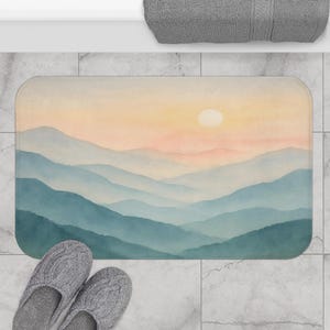 Op de afbeelding: Een rechthoekige badmat met een aquarel landschap van gelaagde blauwe bergen onder een perzik- en gele lucht met een witte zon. Grijze kabelgebreide pantoffels staan op de voorgrond.