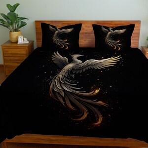 Könnte beinhalten: Schwarzes Bettwäsche-Set mit einem Phönix-Design in Gold und Weiß. Das Set beinhaltet einen Bettbezug und zwei Kissenbezüge, die jeweils das gleiche Vogelmotiv zeigen. Der Phönix wird mit ausgebreiteten Flügeln vor dunklem Hintergrund dargestellt.