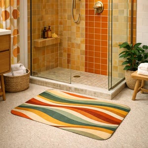 Peut inclure: Un tapis de bain rectangulaire avec un motif de vagues rétro dans les tons orange, vert, jaune et crème. Le tapis est placé devant une douche avec des carreaux orange et beige. Un panier en osier et un petit tabouret en bois sont également visibles.