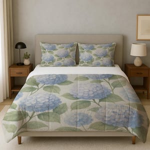 Puede incluir: Una cama con un juego de ropa de cama con estampado de hortensias. El juego incluye un edredón y dos fundas de almohada. El edredón y las fundas de almohada presentan un diseño de flor de hortensia azul claro con hojas verdes sobre un fondo crema.