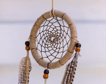 May include: A handcrafted dreamcatcher with a woven web design. The circular frame is wrapped in tan twine, adorned with wooden beads and speckled feathers. The dreamcatcher is suspended by a thin, natural-colored string.