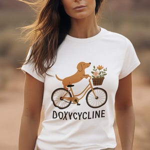 Könnte beinhalten: Ein weißes Kurzarm-T-Shirt mit einer Grafik eines braunen Dackels auf einem Fahrrad mit einem Blumenkorb. Der Text DOXYCYCLINE ist in schwarzen Großbuchstaben unter der Fahrradgrafik auf der Vorderseite des T-Shirts aufgedruckt.