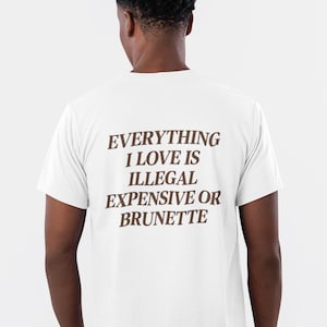 Può includere: T-shirt bianco con la scritta "EVERYTHING I LOVE IS ILLEGAL EXPENSIVE OR BRUNETTE" in lettere marroni. La persona indossa jeans neri e scarpe da ginnastica grigie. L'immagine è ripresa da dietro.