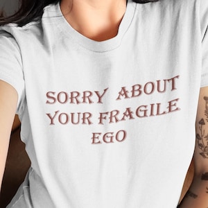 Può includere: Una t-shirt bianca a maniche corte con il testo SORRY ABOUT YOUR FRAGILE EGO stampato in un carattere serif marrone scuro sul petto. Il capo presenta un classico girocollo e una vestibilità standard, offrendo un aspetto pulito e semplice.