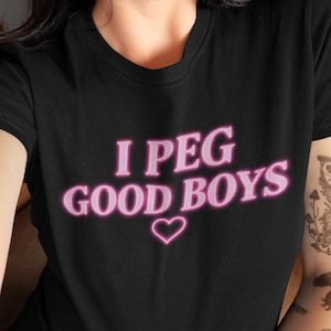 Op de afbeelding: Een zwart t-shirt met korte mouwen met de tekst "I PEG GOOD BOYS" gedrukt in een roze neon-stijl lettertype, vergezeld van een kleine roze hartomtrek gecentreerd direct onder de tekst op de voorkant van het kledingstuk.