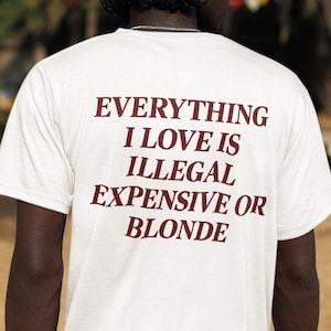 Può includere: Una t-shirt bianca a maniche corte con testo rosso scuro sul retro che recita: EVERYTHING I LOVE IS ILLEGAL EXPENSIVE OR BLONDE. La maglia è abbinata a pantaloncini cargo neri, mostrando uno stile casual con una stampa grafica ben definita.