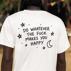 Op de afbeelding: Een wit T-shirt met korte mouwen en zwarte tekst op de achterkant met de tekst "DO WHATEVER THE FUCK MAKES YOU HAPPY", omringd door kleine handgetekende sterren en een maansikkel in zwarte inkt op de bovenrug.