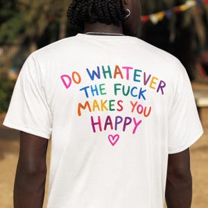 Pode incluir: Uma t-shirt branca de manga curta com o texto multicolorido DO WHATEVER THE FUCK MAKES YOU HAPPY impresso nas costas, acompanhado por um pequeno coração rosa centrado abaixo do texto. A t-shirt é usada com calções cargo pretos.