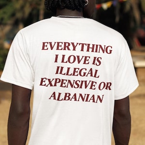 Può includere: Una t-shirt bianca a maniche corte con una scritta in rosso scuro sul retro che recita: EVERYTHING I LOVE IS ILLEGAL EXPENSIVE OR ALBANIAN. Il capo è abbinato a pantaloncini cargo neri con tasche laterali visibili e un bracciale a catena argentato.