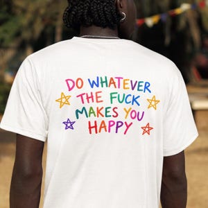 Può includere: Una t-shirt bianca a maniche corte con il testo "DO WHATEVER THE FUCK MAKES YOU HAPPY" stampato in lettere multicolori sul retro. Il design include quattro stelle disegnate a mano nei colori giallo, arancione, viola e rosso.