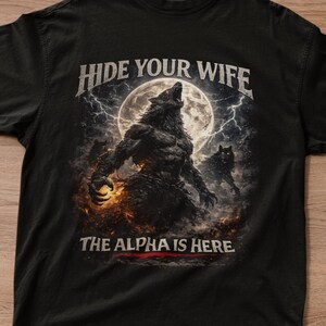 Puede incluir: Camiseta negra con una ilustración detallada de un lobo aullando a la luna, con el texto "HIDE YOUR WIFE" y "THE ALPHA IS HERE". El diseño está sobre un cielo tormentoso.