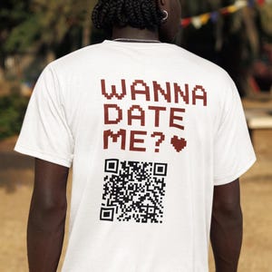 May include: A white short-sleeved t-shirt featuring the dark red pixelated text WANNA DATE ME? followed by a small heart symbol and a black and white QR code printed on the back. The shirt is worn with black cargo shorts and a silver chain bracelet.