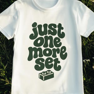 Könnte beinhalten: Ein weißes T-Shirt mit kurzen Ärmeln und dem dunkelgrünen Schriftzug "just one more set" in einer abgerundeten Retro-Schriftart, platziert über einer grafischen Abbildung eines einzelnen rechteckigen Bausteins mit zwei Noppen.