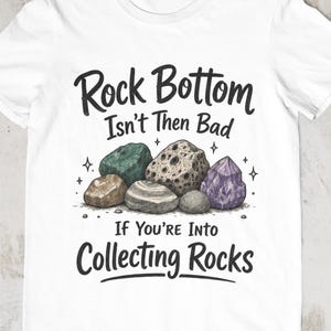 Könnte beinhalten: Ein weißes Kurzarm-T-Shirt mit einer Grafik aus verschiedenen Steinen und Kristallen in Grün-, Braun-, Grau- und Violetttönen. Der Text lautet "Rock Bottom Isn't Then Bad If You're Into Collecting Rocks" in einer schwarzen Schrift.
