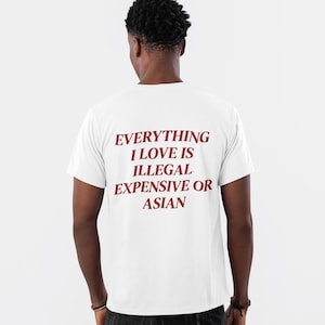 Può includere: T-shirt bianco con la scritta bordeaux "EVERYTHING I LOVE IS ILLEGAL EXPENSIVE OR ASIAN". La persona indossa jeans neri e scarpe da ginnastica grigie. L'immagine è ripresa da dietro.