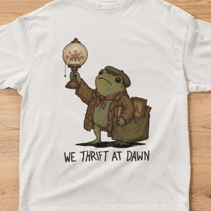 Puede incluir: Una camiseta blanca de manga corta con el gráfico de una rana verde que lleva una chaqueta marrón, un pañuelo rojo y una boina. La rana sostiene una lámpara antigua y lleva una bolsa llena de objetos. El texto WE THRIFT AT DAWN está impreso debajo.