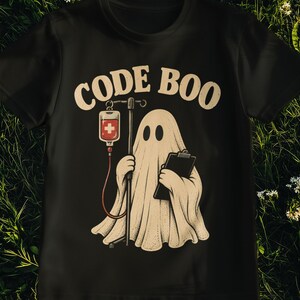 Può includere: Una t-shirt nera con la grafica di un fantasma bianco che tiene in mano una cartellina e un'asta per flebo con una sacca medica rossa. Il testo "CODE BOO" è stampato in un carattere beige arcuato sopra l'illustrazione del fantasma.