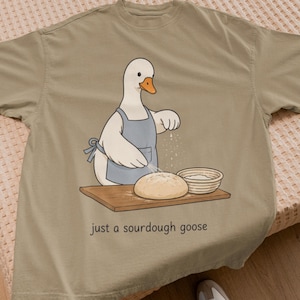 Può includere: Una t-shirt verde oliva chiaro con la grafica di un'oca bianca che indossa un grembiule blu, intenta a spolverare di farina una pagnotta rotonda di pane a lievitazione naturale su un tagliere di legno, con la scritta "just a sourdough goose".