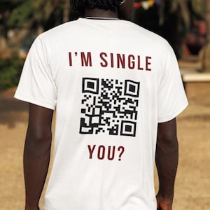 Può includere: Una t-shirt bianca a maniche corte con il testo rosso scuro I'M SINGLE in alto e YOU? in basso, con un grande codice QR in bianco e nero stampato al centro della schiena. La maglietta è indossata con pantaloncini cargo di colore nero.