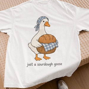 Può includere: Una t-shirt bianca con un'illustrazione grafica di un'oca bianca che indossa un foulard blu a pois bianchi, che trasporta un cesto di vimini marrone con un panno a quadri blu e bianchi contenente una pagnotta, con il testo "just a sourdough goose" stampato sotto.