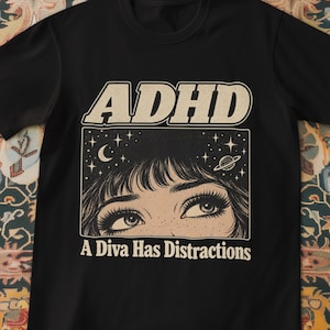 Può includere: Una t-shirt nera a maniche corte con una stampa grafica color crema. Il design include il testo ADHD in alto e A Diva Has Distractions in basso, che incorniciano un'illustrazione dettagliata di occhi rivolti verso un cielo stellato.