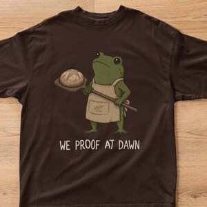 Può includere: Una t-shirt a maniche corte marrone scuro con la grafica di una rana verde che indossa un grembiule beige e tiene in mano una pala di legno con una pagnotta rotonda. Sotto la rana, il testo "WE PROOF AT DAWN" è scritto in un carattere chiaro.