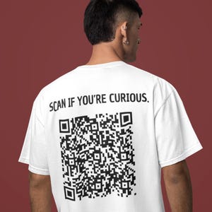 Op de afbeelding: Wit T-shirt met een grote zwarte QR-code en de tekst "SCAN IF YOU'RE CURIOUS." Het T-shirt heeft een ronde hals en is geschikt voor dagelijks gebruik. Het ontwerp is gedrukt op de achterkant van het shirt.