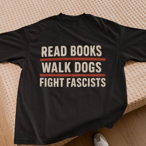 Könnte beinhalten: Ein schwarzes kurzärmeliges T-Shirt mit dem Text READ BOOKS, WALK DOGS, FIGHT FASCISTS in cremeweißen Großbuchstaben, getrennt durch zwei horizontale rote Linien, die mittig auf der Brust des Kleidungsstücks platziert sind.