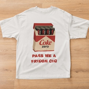 Könnte beinhalten: Weißes T-Shirt mit einer Grafik einer Coke Zero-Schachtel mit Coke Zero-Dosen. Der Text "PASS ME A FRIDGE CIG" ist darunter in Rot gedruckt. Das Shirt besteht aus einem weichen Material.