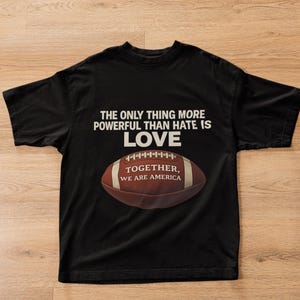 Può includere: T-shirt nero con la scritta "THE ONLY THING MORE POWERFUL THAN HATE IS LOVE" e una grafica di football americano. La grafica del football americano riporta la scritta "TOGETHER, WE ARE AMERICA".