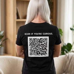 Può includere: T-shirt nera con la scritta "SCAN IF YOU'RE CURIOUS." sopra un grande codice QR. La maglietta è indossata da una persona con lunghi capelli lisci e chiari. La persona è in piedi davanti a uno sfondo neutro.