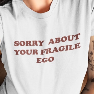 Può includere: Una t-shirt bianca a maniche corte con il testo "SORRY ABOUT YOUR FRAGILE EGO" stampato in un carattere sans-serif marrone scuro sul petto. Il capo presenta un classico girocollo e una vestibilità standard per un look pulito ed essenziale.