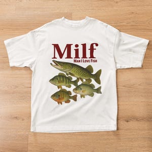 Può includere: T-shirt bianca con una grafica di quattro pesci e la parola "Milf" in bordeaux, con la scritta "Man I Love Fish" sotto. I pesci sono in tonalità di verde, marrone e arancione.